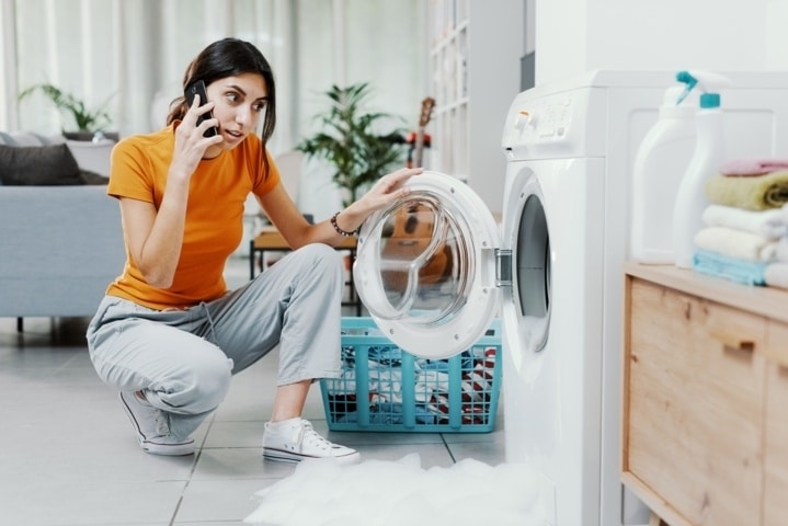Mujer hablando por el móvil junto a una lavadora dañada, preguntándose quién paga las reparaciones en un piso de alquiler